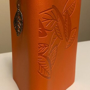 May include: An orange leatherette pen holder with embossed leaf design. A silver leaf charm hangs from a brown leather cord attached to the top. The pen holder has a riveted closure at the top and bottom.