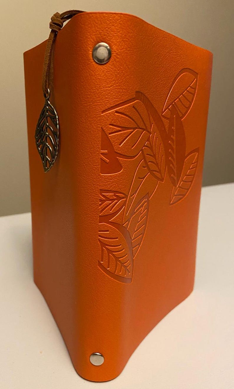 May include: An orange leatherette pen holder with embossed leaf design. A silver leaf charm hangs from a brown leather cord attached to the top. The pen holder has a riveted closure at the top and bottom.