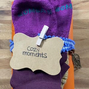 May include: A gift set featuring purple socks with teal text, a brown tag with "Cozy moments" written on it, and a blue and white ribbon. The set is wrapped around an orange item and includes a decorative leaf charm.