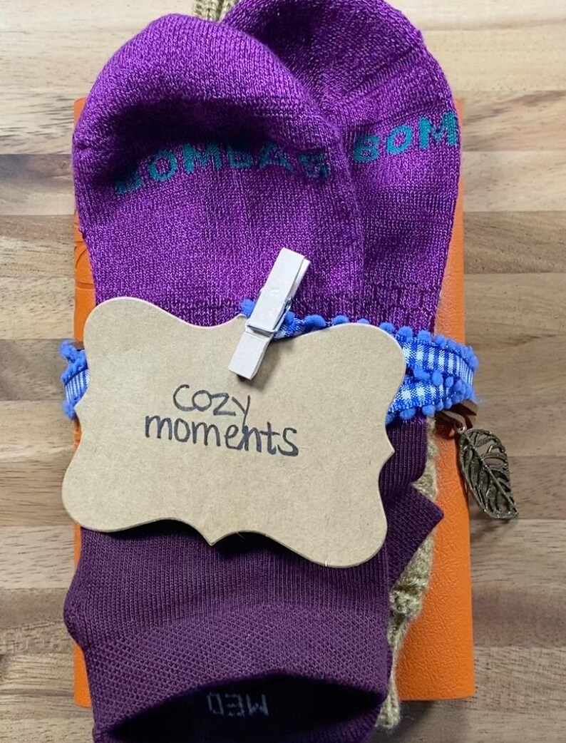 May include: A gift set featuring purple socks with teal text, a brown tag with "Cozy moments" written on it, and a blue and white ribbon. The set is wrapped around an orange item and includes a decorative leaf charm.
