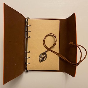 May include: An open, brown leather journal with a leaf charm. The journal has a ring binder and a tan paper interior. The cover is brown with orange edges. A brown leather cord with a metal leaf charm rests on the open pages.
