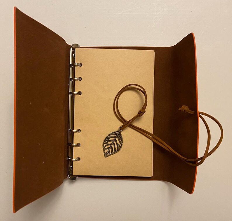 May include: An open, brown leather journal with a leaf charm. The journal has a ring binder and a tan paper interior. The cover is brown with orange edges. A brown leather cord with a metal leaf charm rests on the open pages.