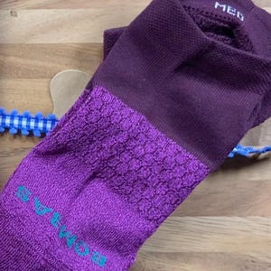 May include: A purple sock with a textured toe and heel, and the word "MED" printed on the cuff. The sock has a darker purple cuff and a lighter purple toe and heel. The brand name is printed in teal on the toe.