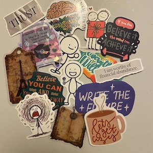 May include: A collection of colorful stickers with various designs and text. Stickers include phrases like "TRUST", "Believe you can", and "Let's get cozy". Other stickers feature illustrations of a brain, a coffee mug, and stick figures.