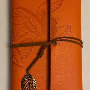 May include: Orange leather journal with embossed leaf design. The journal is secured with a brown cord and a decorative leaf charm. The cover features a textured surface and a flap closure.