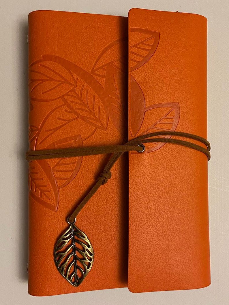 May include: Orange leather journal with embossed leaf design. The journal is secured with a brown cord and a decorative leaf charm. The cover features a textured surface and a flap closure.