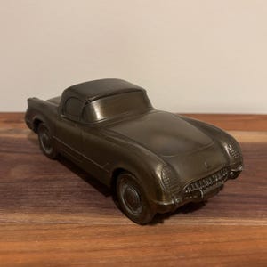 May include: A bronze-colored vintage convertible car model. The car has a sleek design with a hardtop, detailed headlights, and a front grill. The model is displayed on a wooden surface.