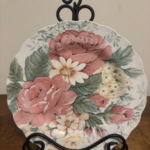 May include: A decorative plate with a scalloped edge, featuring a floral design of pink roses, yellow flowers, and green leaves. The plate is displayed on a black wrought iron stand, showcasing its intricate details and colors.