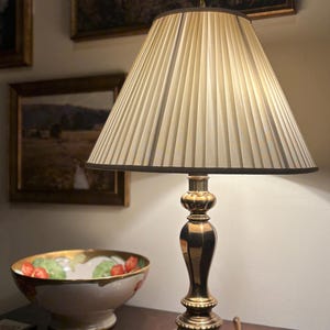 May include: A brass table lamp with a pleated lampshade. The lamp has a decorative base and a curved handle. A decorative bowl with a floral design is also visible.