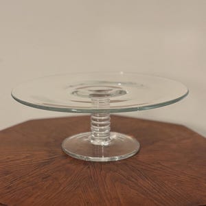 May include: A clear glass cake stand with a circular top and a ribbed stem. The stand is displayed on a dark wooden table. The cake stand is a classic design, suitable for displaying desserts or other items.