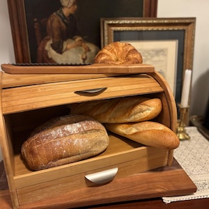 May include: A wooden bread box with a roll-top lid, holding various loaves of bread and a croissant. The box is made of light-colored wood and has a dark wooden base. A bread knife rests on top.