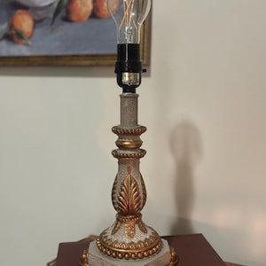 May include: A decorative table lamp with a gold and white ornate base and a clear lightbulb. The lamp has a crackle finish and a leaf design. The lamp is on a dark red surface.