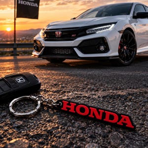 May include: A black Honda key fob with a metal ring attached to a rectangular black and red rubber keychain featuring the word HONDA in bold red letters. The keychain rests on a textured asphalt surface with a white Honda car in the background.
