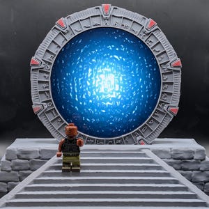 May include: A gray, circular Stargate model with red accents and a bright blue energy field in the center. A Lego figure stands on a gray stone staircase in front of the Stargate.