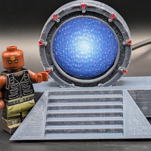 May include: A gray 3D-printed circular stargate model with a glowing blue center and red accents, mounted on a base with a set of stairs. Beside the structure stands a small brown-skinned plastic minifigure wearing a black vest and tan pants.