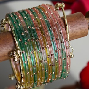 May include: A collection of vibrant bangles in green, yellow, and pink, each decorated with tiny, glittering studs. The bangles are displayed on a wooden dowel, featuring gold-toned metal clasps and details. The background is blurred, with a mirror and red flowers visible.