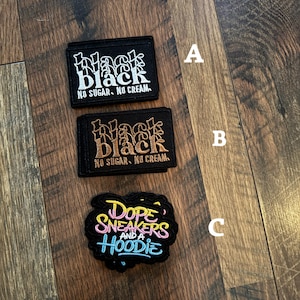 May include: Three embroidered patches on a wooden surface. Two rectangular patches feature the word "Black" in stylized lettering, with "NO SUGAR. NO CREAM." below. The third patch is oval-shaped, with the words "DOPE SNEAKERS AND A HOODIE."
