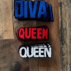 May include: Three embroidered patches with the words "DIVA" in blue, "QUEEN" in red, and "QUEEN" in white. Each patch has a black border and a textured, raised appearance. The patches are displayed on a wooden surface.