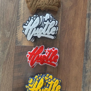 May include: Four embroidered patches spelling the word "Hustle" in a script font. The patches are in brown, white with black trim, red with white trim, and yellow with blue trim. The patches are on a wooden surface.
