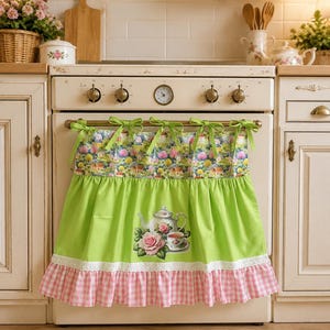 Può includere: Una tendina per forno con un pannello superiore a stampa paesaggistica colorata, una sezione centrale verde lime con una grafica di una teiera, una tazzina e una rosa, rifinita con un bordo in pizzo bianco e una balza a quadretti rosa e bianchi.