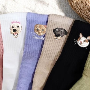 Könnte beinhalten: Eine Auswahl an farbigen Rippstricksocken, jede mit dem Gesicht eines anderen Haustiers bestickt. Die Socken sind in den Farbtönen Rosa, Weiß, Lavendel, Beige, Schwarz und Olivgrün gehalten. Einige Socken haben Haustiernamen unter dem Gesicht des Haustiers.