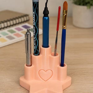 May include: A peach-colored, star-shaped desk organizer with a heart embossed on the front. It features five cylindrical slots holding a silver pen, a black acrylic paint marker, a blue tool, a thin red tool, and a paintbrush with a blue handle.