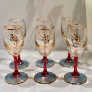 Puede incluir: Juego de seis copas de vino vintage. Cada copa presenta un cuenco transparente con borde dorado y diseño de rosa dorada. El tallo es rojo y la base azul claro con ribete dorado. Las copas están sobre una superficie blanca.