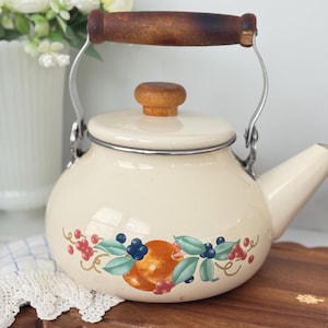 Puede incluir: Tetera de esmalte color crema con asa y pomo de tapa de madera. La tetera presenta un diseño decorativo de frutas y hojas en tonos naranja, azul y rojo. La boquilla está en el lado derecho. Un artículo de cocina encantador.