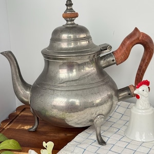 May include: A vintage pewter teapot with a wooden handle and finial. The teapot has a classic design with a curved spout and sits on three small feet. A white ceramic chicken-shaped bell is next to the teapot.