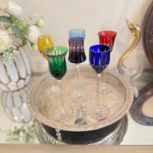 Puede incluir: Seis copas de vino de colores, amarillo, verde, morado, azul y rojo, dispuestas sobre una bandeja plateada. Las copas tienen un diseño tallado y están sobre una superficie reflectante. La bandeja es redonda.