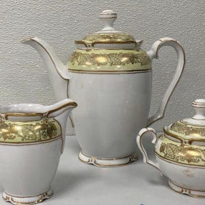 May include: A white porcelain tea set consisting of a teapot, a creamer, and a lidded sugar bowl. Each piece features a pale yellow band around the top decorated with a gold leaf and vine pattern, accented by gold trim on the rims and handles.