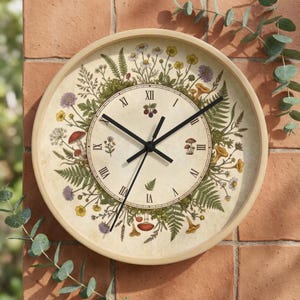 Puede incluir: Reloj de pared redondo con marco de madera clara. La esfera del reloj presenta un diseño botánico con helechos, setas y flores silvestres. Las manecillas negras indican la hora. Números romanos.
