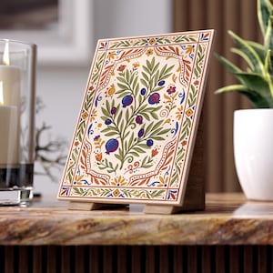 May include: A rectangular decorative tile with a floral design, featuring a central tree with purple fruit and green leaves. The tile is framed with a border of orange, blue, and yellow accents, and is propped up on a wooden stand.