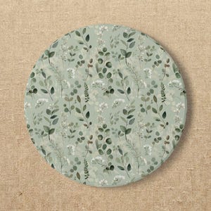 May include: A circular coaster featuring a sage green background with a repeating pattern of dark green eucalyptus leaves, thin stems, and small white flower clusters. The design is printed across the entire surface of the round object.