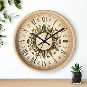 Puede incluir: Reloj de pared redondo con marco de madera clara y esfera crema. La esfera del reloj presenta números romanos y un diseño botánico central con setas, helechos y flores. Las manecillas negras indican la hora.