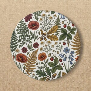 Puede incluir: Un posavasos circular con una ilustración botánica sobre fondo blanco. El diseño incluye amapolas rojas, flores de manzanilla blancas, campanillas azules, helechos verdes, hojas de roble doradas con bellotas y pequeñas fresas rojas con hojas verdes.