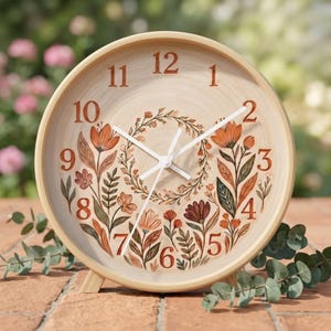 May include: A round analog clock with a light wood frame and a beige face. The clock features dark orange numerals from 1 to 12 and a central floral illustration with orange, brown, and green leaves and flowers. It has three white clock hands.