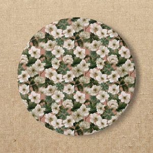 Puede incluir: Un posavasos circular con un patrón botánico repetitivo de flores blancas con centros amarillos y hojas de color verde oscuro sobre un fondo rosa empolvado. El diseño cubre toda la superficie del objeto redondo.