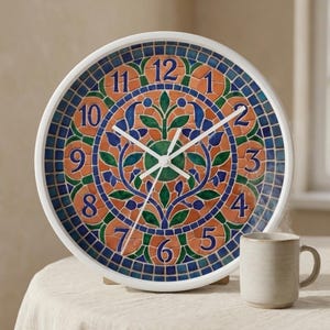 Peut inclure: Une horloge murale ronde avec un cadre blanc et un cadran de style mosaïque. Le cadran présente un motif coloré avec des éléments orange, bleus et verts. Les aiguilles sont blanches et les chiffres bleus. Une tasse beige est à droite.