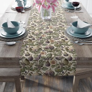 May include: A beige table runner with a fig and leaf pattern. The design features figs in shades of purple and brown, surrounded by green leaves and small purple berries. The runner is set on a wooden table with place settings.