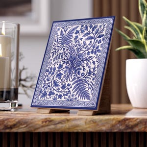 Könnte beinhalten: Eine rechteckige Keramikfliese mit einer detaillierten blau-weißen botanischen Illustration von Farnen, Efeu und Blumen. Die Fliese ist von einem dekorativen Rand umgeben und wird auf einem kleinen Holzständer aufrecht präsentiert.