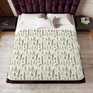 May include: A cream-colored blanket with a repeating pattern of gray fern illustrations. The blanket is on a bed with white pillows and a dark purple velvet headboard. The bed is flanked by two black nightstands.