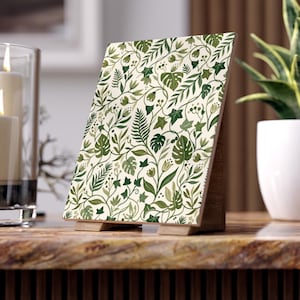 May include: A rectangular tabletop sign featuring a botanical pattern of green leaves, ferns, and small flowers on a cream background. The sign is supported by a light-colored wooden easel stand, displaying a dense, repeating nature-inspired illustration.