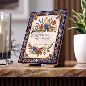 May include: A decorative ceramic plaque with a floral design and the words "GRACE & PEACE EST. 2025" in navy blue. The plaque features a navy blue border with orange and gold accents. It is propped up on a wooden stand.