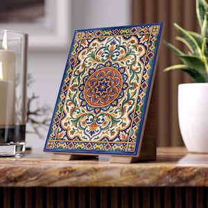 Puede incluir: Una baldosa decorativa de cerámica con un diseño intrincado. La baldosa presenta un medallón central con un patrón de estrella en tonos naranja y azul, rodeado de motivos florales y geométricos en verde, amarillo y azul. Se muestra sobre un soporte de madera.