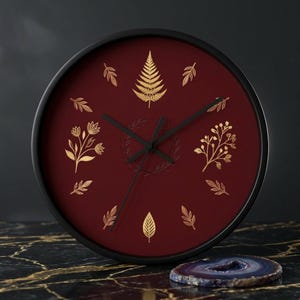 May include: Round wall clock with a black frame and a deep red face. The clock face features gold-colored botanical designs, including leaves and floral patterns. The clock hands are black.