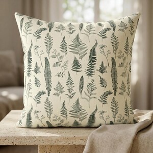 Peut inclure: Coussin carré avec un fond crème et un motif répété d'illustrations de fougères vertes. Le coussin est posé sur une table de couleur claire, suggérant un thème de décoration intérieure.