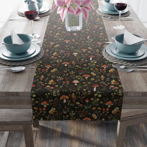 May include: A dark table runner with a repeating pattern of mushrooms, flowers, and foliage in shades of red, orange, yellow, and green. The runner is set on a wooden table with place settings, wine glasses, and a vase of pink lilies.