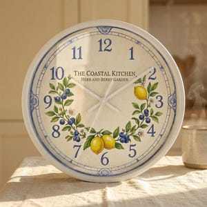 May include: A round wall clock with a white face and a blue rim. The clock features a decorative design of lemons, blueberries, and greenery. The text "THE COASTAL KITCHEN, HERB AND BERRY GARDEN" is printed on the clock face.