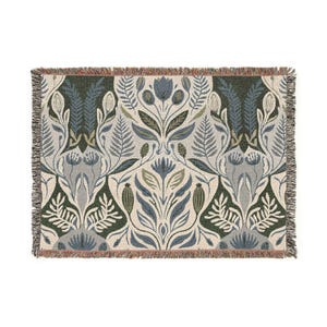 May include: A rectangular woven throw blanket featuring a symmetrical botanical pattern of blue, sage green, and dark forest green flowers and ferns on a cream background, finished with a short, multi-coloured fringed edge on all four sides.